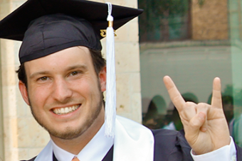 UT student in cap and gown holding up the hook em sign
